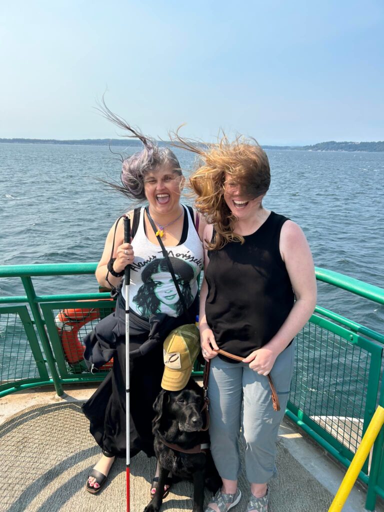 2 women on a ferry with their hair flying wild in the breeze and one with a white cane, one with a black guide dog.